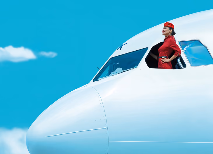 Flight attendant in red uniform standing confidently in an airplane cockpit window against a blue sky.