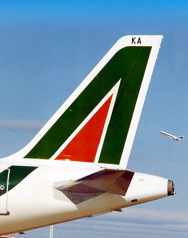 Tail of an Alitalia airplane with green and red logo against blue sky and another plane flying in the distance.